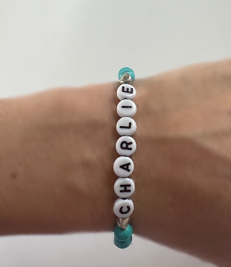 Op de afbeelding: Een witte kralen armband met de naam "CHARLIE" gespeld in zwarte letters. De kralen worden gescheiden door turquoise kralen.