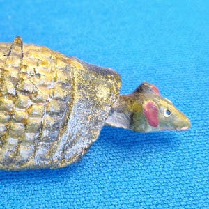 May include: A vintage, hand-painted ornament in the shape of a mouse riding on a turtle. The turtle is primarily gold and brown, with a textured shell. The mouse has a green head with red ears. A small metal loop is attached for hanging.