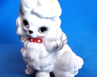 Könnte beinhalten: Eine weiße Keramik-Pudel-Figur mit einem roten Kragen mit weißen Punkten. Der Pudel hat schwarze Augen, rosa Wangen und einen gelben Fleck auf dem Kopf. Die Figur sitzt.