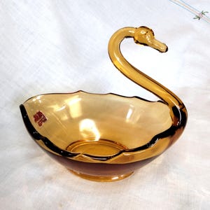 May include: Amber-colored glass swan-shaped bowl. The bowl has a scalloped rim and a dark brown base. The swan's neck curves gracefully, forming a handle. A small label is visible on the side. This decorative piece is suitable for serving or display.