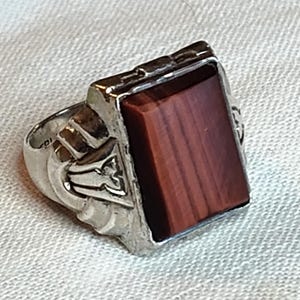 May include: A silver-toned ring featuring a rectangular, reddish-brown stone with a smooth, flat surface. The metal band displays intricate, raised geometric detailing along the sides, framing the central stone in a structured, metallic setting.