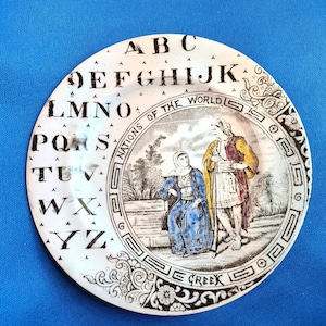 May include: A white ceramic plate with a black border and a colorful image of a man and woman in traditional Greek clothing. The plate has the alphabet printed around the edge and the words "Nations of the World" and "Greek" printed on the bottom.