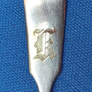 May include: A silver-toned metal spoon handle featuring a polished finish and a distinct, engraved gothic-style letter K centered on the flat surface. The top of the handle has a subtle, rounded, heart-shaped indentation near the edge.