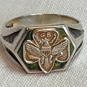 May include: A silver-toned ring featuring a hexagonal face with a green enamel background. Centered on the face is a gold-toned emblem of an eagle with spread wings, a shield on its chest, and the letters GS above the eagle's head.