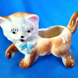 May include: A ceramic planter shaped like a brown and white cat with a blue bow. The cat is walking and has a small pot on its back.