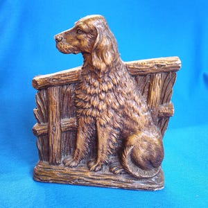 May include: A brown, molded resin bookend featuring a relief sculpture of a long-haired dog sitting in profile against a textured wooden fence background. The dog has detailed fur, a pointed snout, and floppy ears, with a wood-grain base.