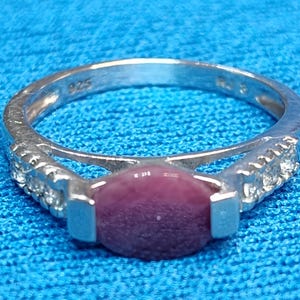 May include: A silver ring featuring a central oval-shaped dark pink stone flanked by three small clear stones on each side of the band. The inner band is clearly stamped with the text 925, indicating sterling silver composition for this jewelry piece.