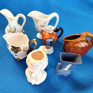 May include: A collection of six small ceramic pitchers and vases in various shapes and colors, including white with gold accents, a miniature orange teapot with a landscape scene, a textured brown pitcher, a white bust vase, and a blue relief vase.