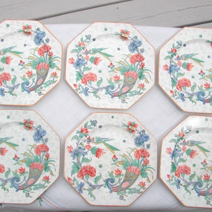 May include: Six octagonal plates with a floral design. Each plate features a central arrangement of red, blue, and green flowers and foliage, with a bird and horn of plenty motif. The plates have a cream-colored background and a light orange border.