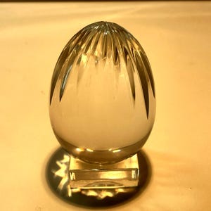 Puede incluir: Un pisapapeles de cristal transparente con forma de huevo que presenta facetas verticales talladas que irradian desde la punta superior. El huevo descansa sobre una base cuadrada de cristal transparente, creando un aspecto liso y brillante.