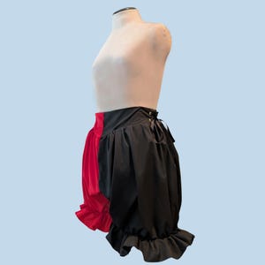 May include: A two-toned, red and black, ruffled skirt displayed on a mannequin. The skirt is split down the middle, with a red panel on the left and a black panel on the right. The skirt has a high waist and a lace-up detail on the side.
