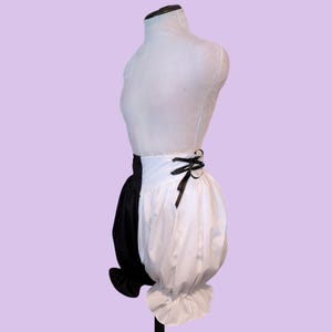 May include: High-waisted bloomers featuring a split color design with one black leg and one white leg. The garment includes a wide white waistband with black ribbon lace-up detailing on the side and ruffled hems at the bottom of each leg.