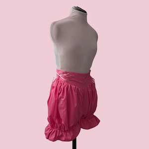 May include: Bright pink bloomers featuring a high-waisted design with a lace-up front closure using matching pink ribbon through metal eyelets. The garment has a voluminous, gathered silhouette with ruffled leg openings, displayed on a mannequin.