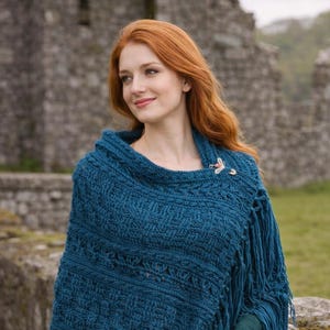May include: A teal blue knitted shawl with fringe. The shawl is fastened with a decorative brooch. The woman is wearing a long-sleeved green shirt and a brown skirt. The background features a stone wall and green grass.
