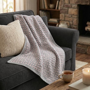 Puede incluir: Una manta de ganchillo gris y rosa sobre un sofá gris oscuro, con un cojín color crema. Una mesa de centro de madera tiene una taza de té y una vela encendida. El fondo incluye una chimenea y una estantería.