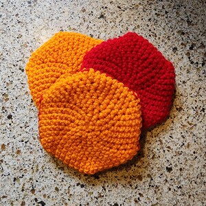 May include: Three crocheted pot holders, one red and two orange, on a speckled countertop.