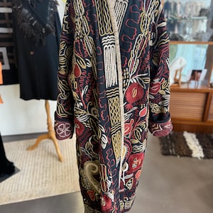 May include: A long black coat with detailed embroidery in red, gold, and white. The coat features a floral and geometric pattern, with a contrasting trim. The sleeves have a patterned cuff. The coat is displayed on a mannequin. The coat is a vintage style.