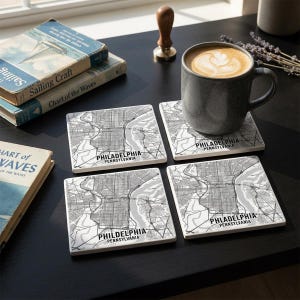 May include: A set of four square coasters featuring a black and white map of Philadelphia, Pennsylvania. Each coaster displays the city street grid, a river, and the text Philadelphia Pennsylvania printed in a bold, black sans-serif font.