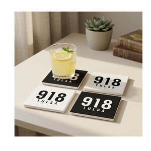 May include: A set of four square coasters featuring a graphic design with the number 918 and the text TULSA. Two coasters have a white background with black text, and two coasters have a black background with white text, arranged on a surface.