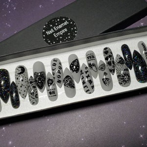May include: A set of 12 white press-on nails with black designs. The designs include a crescent moon, stars, a heart with an eye, a hand with an eye, a skull, a pentagram, and the words "GOOD BYE", "YES", "WITCH", "NOPE", "CODE", and "6661390".