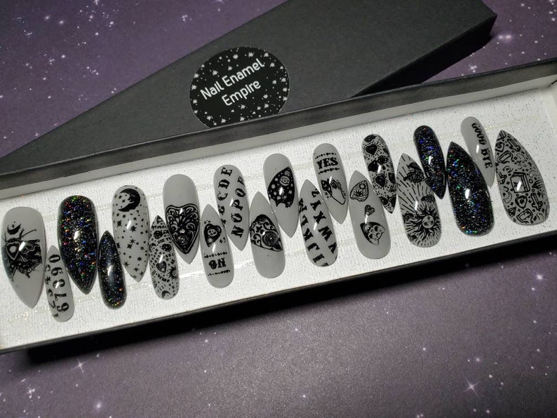 May include: A set of 12 white press-on nails with black designs. The designs include a crescent moon, stars, a heart with an eye, a hand with an eye, a skull, a pentagram, and the words "GOOD BYE", "YES", "WITCH", "NOPE", "CODE", and "6661390".
