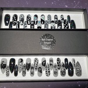 May include: Two black boxes with white text that says "Nail Enamel Empire" on the top box. The boxes are open and contain a set of 20 black and white press-on nails with various designs. The designs include a Ouija board, an eye, a crescent moon, and a heart.