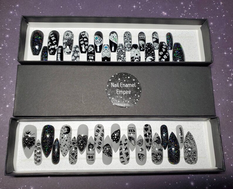 May include: Two black boxes with white text that says "Nail Enamel Empire" on the top box. The boxes are open and contain a set of 20 black and white press-on nails with various designs. The designs include a Ouija board, an eye, a crescent moon, and a heart.