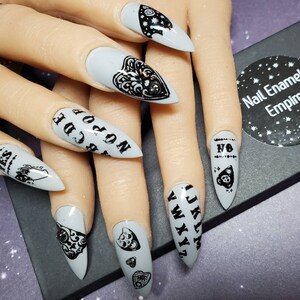 May include: Close-up of a hand with gray nail polish and black nail art featuring a Ouija board, a heart, and a planchette. The nails are long and pointed.