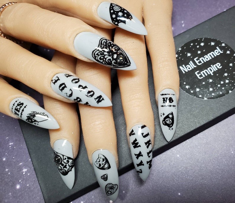 May include: Close-up of a hand with gray nail polish and black nail art featuring a Ouija board, a heart, and a planchette. The nails are long and pointed.
