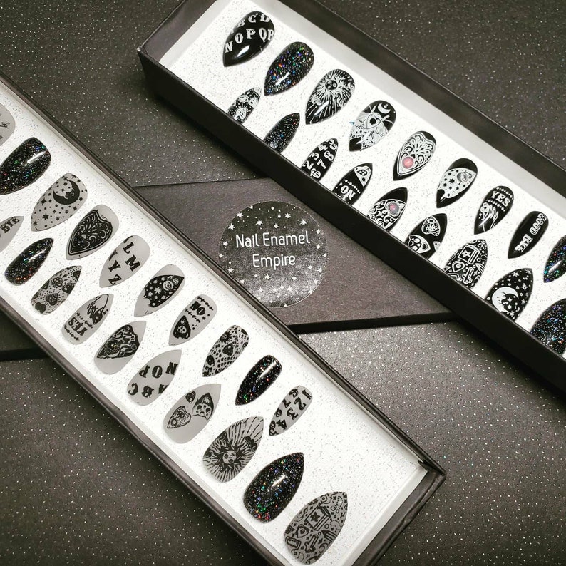 May include: Two black boxes with white trim containing sets of black and white press-on nails with various designs. The designs include a Ouija board, a crescent moon, an eye, and a heart. The boxes are labeled "Nail Enamel Empire".