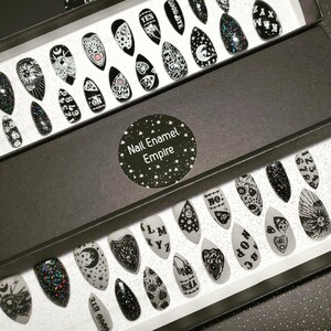 May include: A black box with a white label that says "Nail Enamel Empire" and is filled with black and white nail tips. The nail tips have various designs, including a crescent moon, a heart, a hand with a finger pointing, and a Ouija board.