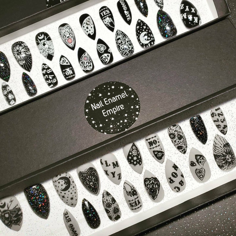 May include: A black box with a white label that says "Nail Enamel Empire" and is filled with black and white nail tips. The nail tips have various designs, including a crescent moon, a heart, a hand with a finger pointing, and a Ouija board.