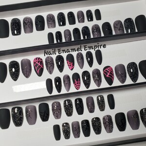 May include: Set of 24 black and gray coffin-shaped false nails with spiderweb designs. Some nails have pink spiderwebs, some have black spiderwebs, and some have black glitter. The nails are arranged in a box with the text "Nail Enamel Empire".