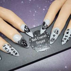 May include: Set of gray stiletto false nails with black designs. The designs include a Ouija board, a heart with an eye, and a bat. The nails are displayed on a black surface with a white sticker that says "Nail Enamel Empire".
