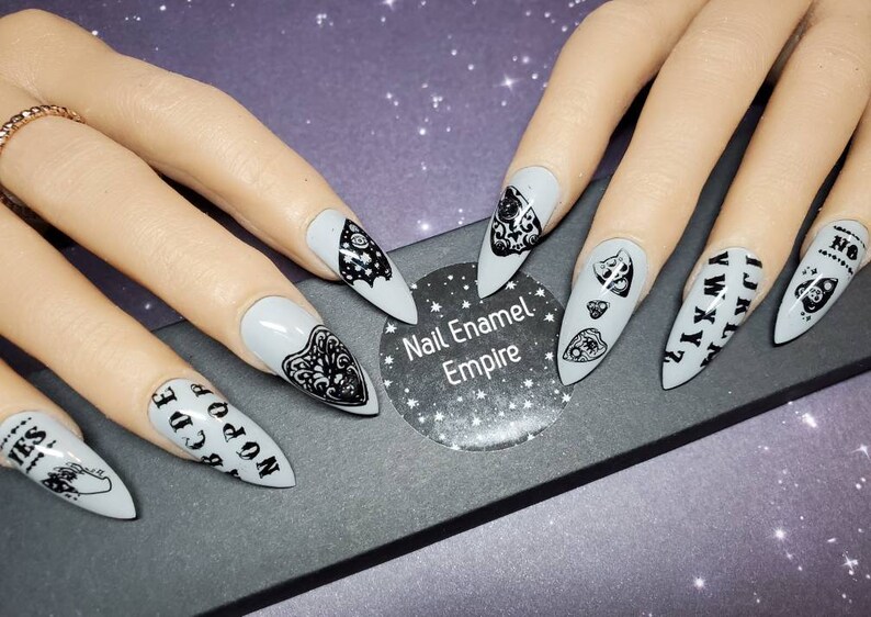 May include: Set of gray stiletto false nails with black designs. The designs include a Ouija board, a heart with an eye, and a bat. The nails are displayed on a black surface with a white sticker that says "Nail Enamel Empire".