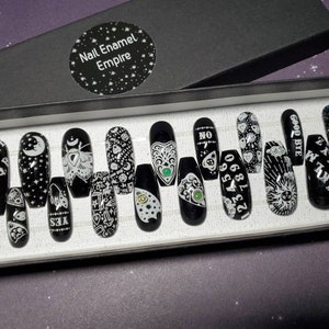 May include: A set of 14 black press-on nails with white designs. The designs include a crescent moon, a moth, a Ouija board, a heart with an eye, a sun, and the words "GOODBYE", "I AM", "YES", "NO", "1234567890", and "CODE", "NOPE", "DEER".