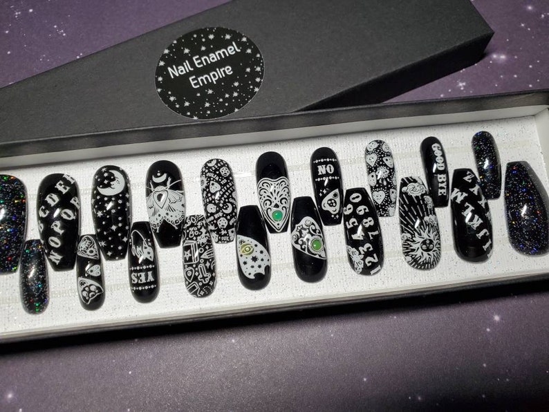 May include: A set of 14 black press-on nails with white designs. The designs include a crescent moon, a moth, a Ouija board, a heart with an eye, a sun, and the words "GOODBYE", "I AM", "YES", "NO", "1234567890", and "CODE", "NOPE", "DEER".