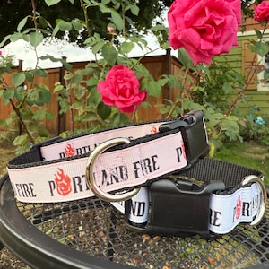 May include: Two pet collars featuring a light pink and white ribbon overlay on black webbing. The ribbon displays the text PORTLAND FIRE in black capital letters with a red flame graphic. Each collar includes a black plastic buckle and a metal D-ring.