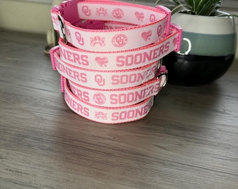 May include: Four pink dog collars with the word "Sooners" and a heart design. The collars are stacked on top of each other.