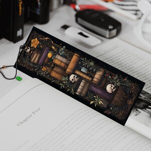 May include: A black bookmark with a colorful illustration of a stack of books, skulls, and plants. The bookmark has a black tassel with a white bead.