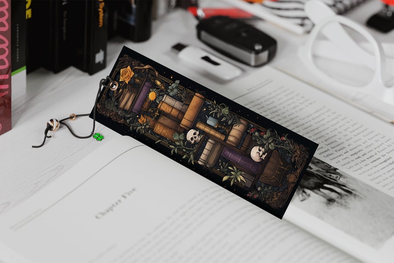 May include: A black bookmark with a colorful illustration of a stack of books, skulls, and plants. The bookmark has a black tassel with a white bead.