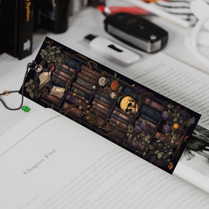 May include: A black bookmark with a detailed illustration of a bookshelf filled with books, vines, flowers, and a skull. The bookmark has a black tassel with a white and black bead.