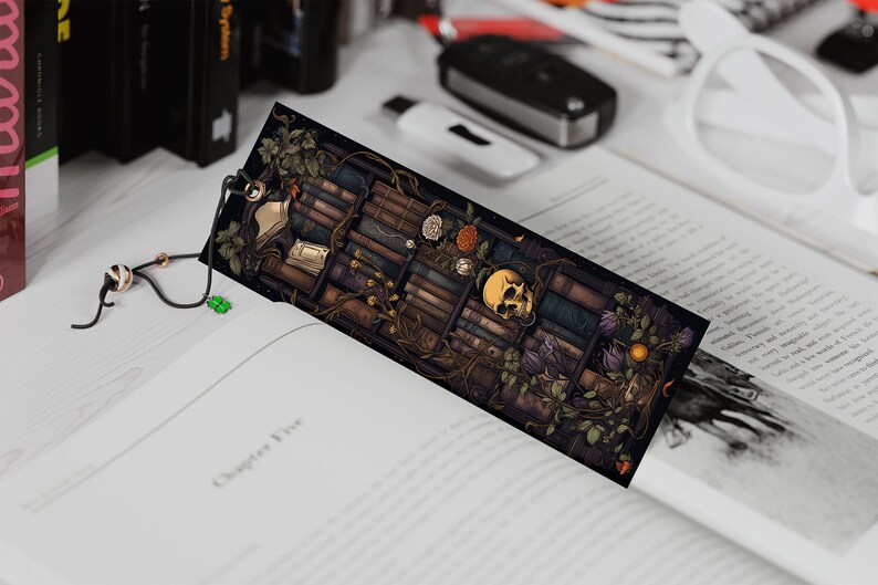 May include: A black bookmark with a detailed illustration of a bookshelf filled with books, vines, flowers, and a skull. The bookmark has a black tassel with a white and black bead.