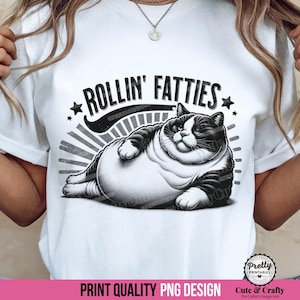 May include: White t-shirt with a black and white illustration of a reclining, plump cat. The text "ROLLIN' FATTIES" is above the cat. The design includes star accents and a sunburst background.