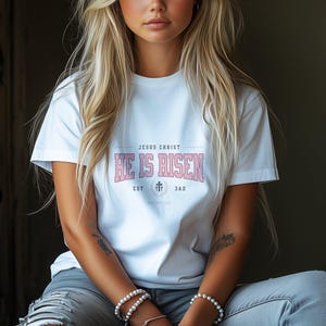May include: White t-shirt with a pink graphic that says "Jesus Christ He is Risen" with a cross and the dates "EST 33 AD".