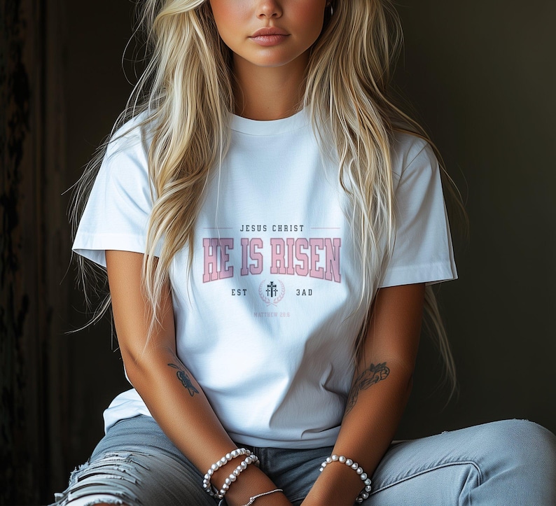 May include: White t-shirt with a pink graphic that says "Jesus Christ He is Risen" with a cross and the dates "EST 33 AD".