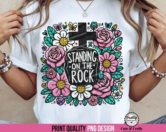May include: White t-shirt featuring a colorful graphic design. The design includes pink roses, daisies, butterflies, and the text "STANDING ON THE ROCK" within a cross-shaped frame. The design is surrounded by green leaves.