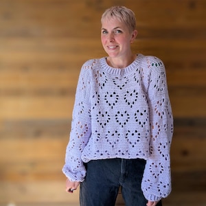 Könnte beinhalten: Ein helllila gestrickter Pullover mit einem Spitzenmuster. Der Pullover hat eine lockere Passform und lange Ärmel.