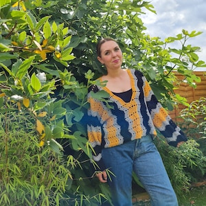 Peut inclure: Une femme portant un haut en crochet jaune et noir avec un col en V et des manches longues. Elle se tient devant un buisson vert feuillu.