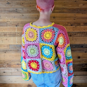 Puede incluir: Un colorido su&eacute;ter de ganchillo con un dise&ntilde;o de patchwork con motivos florales cuadrados. El su&eacute;ter muestra una vibrante paleta de rosa, amarillo, azul y verde, con un escote y pu&ntilde;os festoneados. La prenda se lleva sobre unos vaqueros azules.